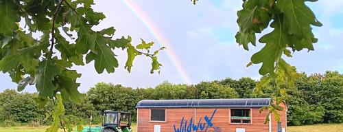 Ein Theaterwagen von WildWux, der an einen Trecker gekoppelt ist, auf einer Wiese mit Regenbogen im Hintergrund. 33d5f93fd09966e61f0a0e1bea18427bd5d026b9